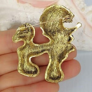 May include: A gold-toned metal brooch shaped like a poodle in a standing pose. The reverse side of the brooch features a textured, brushed gold finish and includes a horizontal pin mechanism with a rotating clasp for secure attachment to fabric.