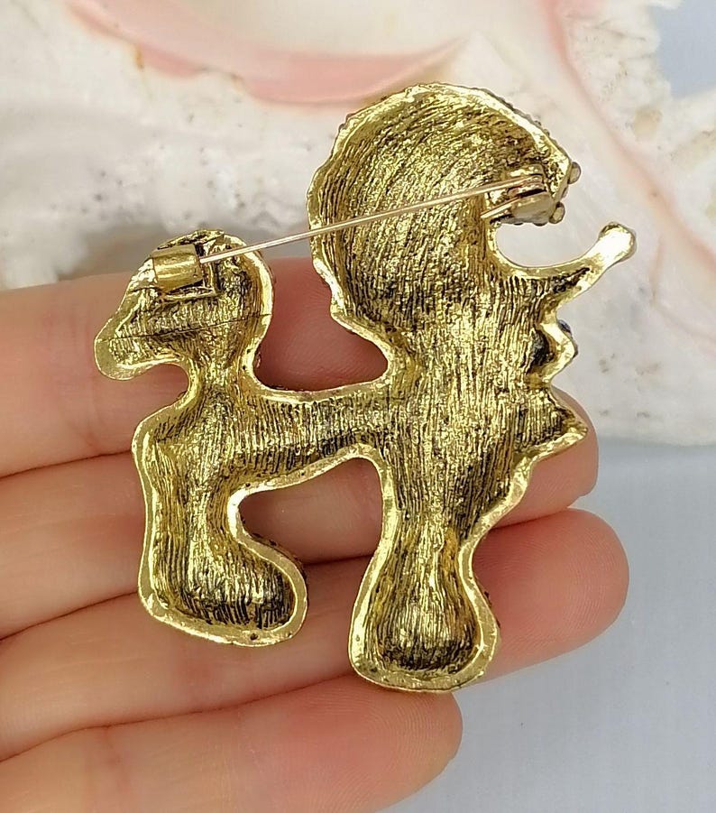 May include: A gold-toned metal brooch shaped like a poodle in a standing pose. The reverse side of the brooch features a textured, brushed gold finish and includes a horizontal pin mechanism with a rotating clasp for secure attachment to fabric.