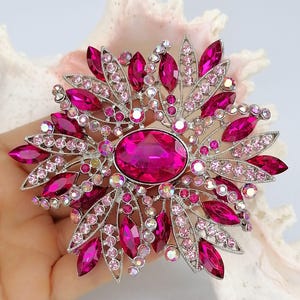 Op de afbeelding: Een zilverkleurige broche met een grote centrale ovale fuchsia kristal, omringd door kleinere marquise-geslepen fuchsia stenen en talloze ronde lichtroze en iriserende kristallen in een stralend, stervormig bloemmotief.