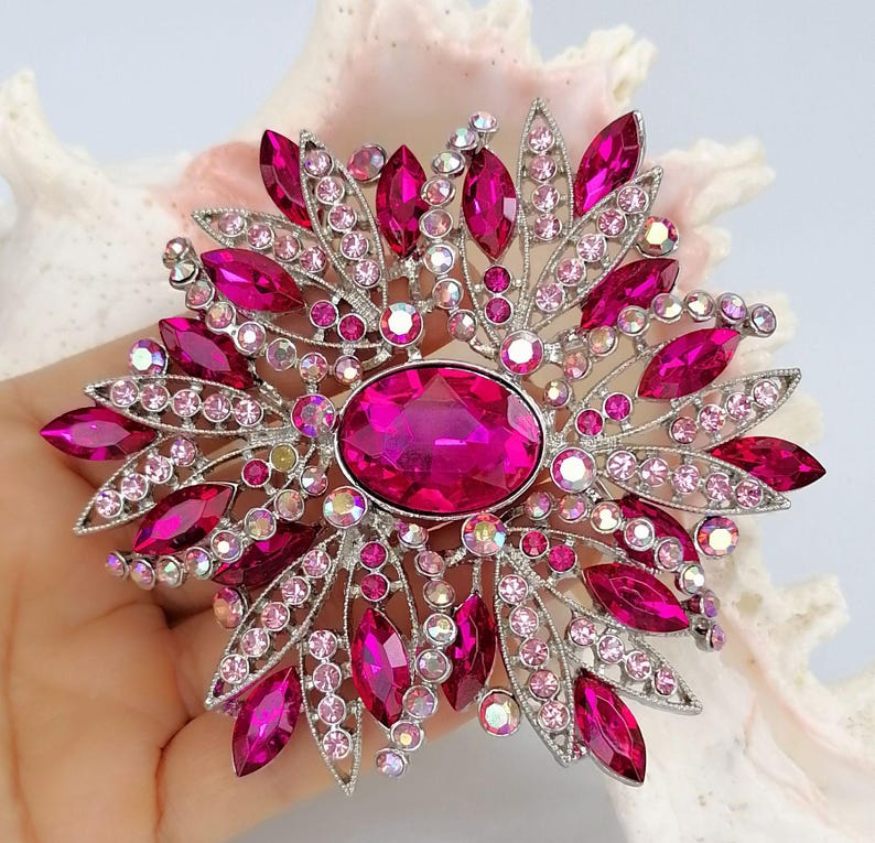Op de afbeelding: Een zilverkleurige broche met een grote centrale ovale fuchsia kristal, omringd door kleinere marquise-geslepen fuchsia stenen en talloze ronde lichtroze en iriserende kristallen in een stralend, stervormig bloemmotief.