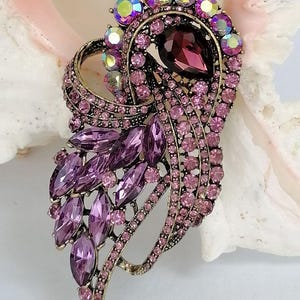 May include: A decorative brooch featuring a dark purple teardrop-shaped center stone surrounded by smaller light purple round crystals and iridescent stones. The metal setting is an antique gold tone with a curved, layered design.