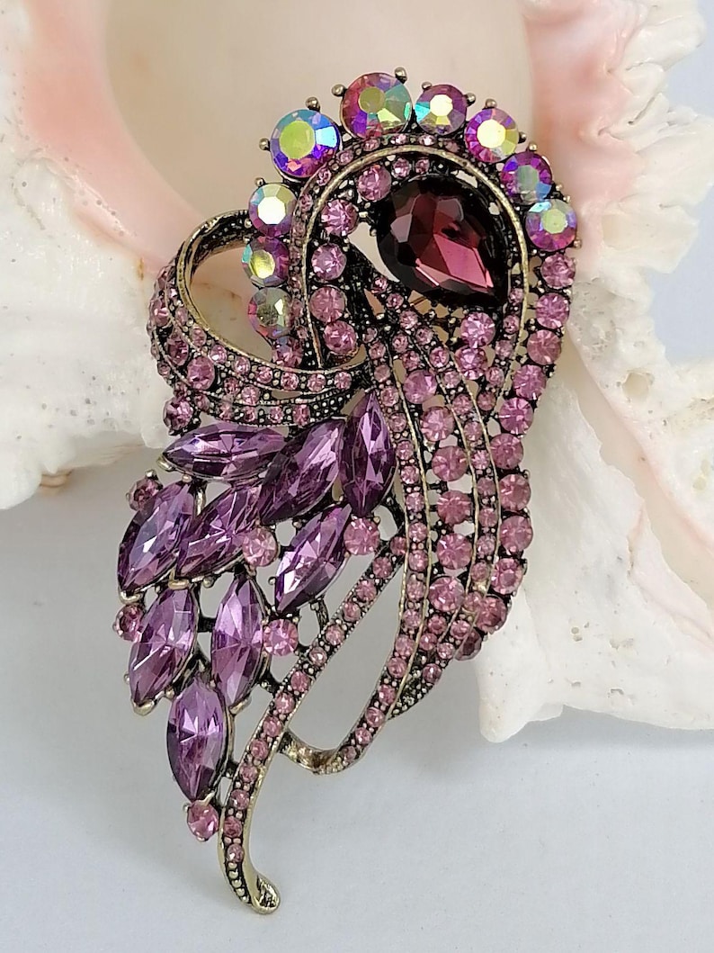 May include: A decorative brooch featuring a dark purple teardrop-shaped center stone surrounded by smaller light purple round crystals and iridescent stones. The metal setting is an antique gold tone with a curved, layered design.