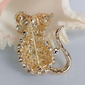 May include: A gold-colored metal brooch in the shape of a mouse. The brooch has a textured surface and a pin on the back for attachment. The mouse has a long, curved tail and detailed features.