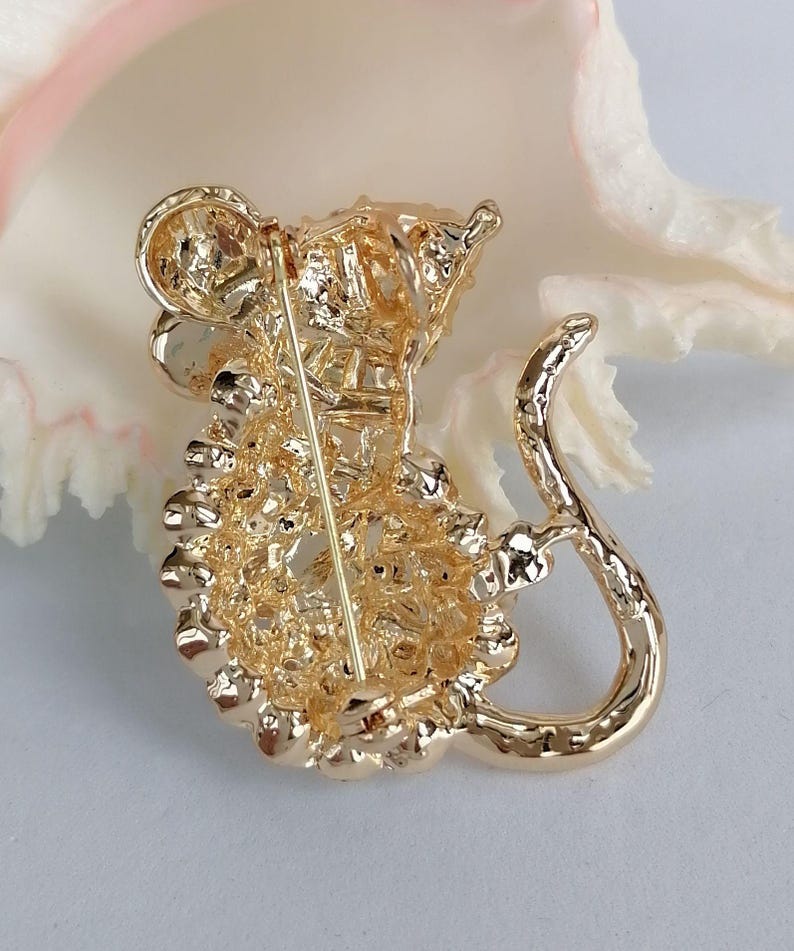 May include: A gold-colored metal brooch in the shape of a mouse. The brooch has a textured surface and a pin on the back for attachment. The mouse has a long, curved tail and detailed features.