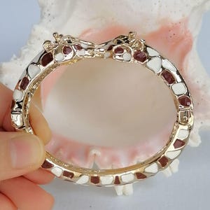May include: A gold-tone hinged bangle bracelet featuring two giraffe heads meeting at the center. The bracelet is decorated with an enamel pattern of white and dark brown patches, mimicking the natural markings of a giraffe's coat throughout.