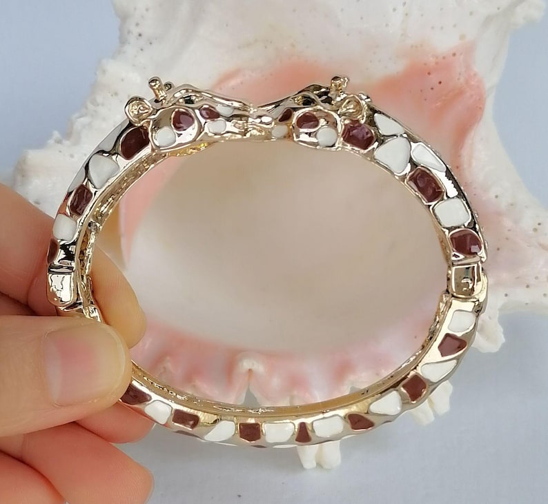 May include: A gold-tone hinged bangle bracelet featuring two giraffe heads meeting at the center. The bracelet is decorated with an enamel pattern of white and dark brown patches, mimicking the natural markings of a giraffe's coat throughout.