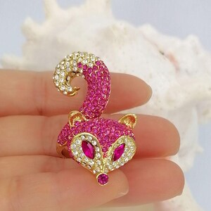 K&ouml;nnte beinhalten: Ein goldfarbener Ring mit einem fuchsf&ouml;rmigen Design, das mit leuchtend rosa und klaren Kristallen besetzt ist. Der Fuchskopf hat zwei marquise-geschliffene rosa Kristallaugen und eine kleine rosa Kristallnase sowie einen geschwungenen Schwanz.
