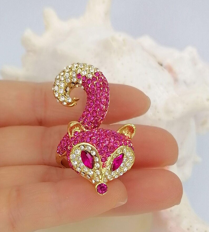 K&ouml;nnte beinhalten: Ein goldfarbener Ring mit einem fuchsf&ouml;rmigen Design, das mit leuchtend rosa und klaren Kristallen besetzt ist. Der Fuchskopf hat zwei marquise-geschliffene rosa Kristallaugen und eine kleine rosa Kristallnase sowie einen geschwungenen Schwanz.