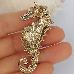 May include: A gold-toned seahorse brooch with a textured surface and a pin closure. The seahorse has a detailed design, including a curled tail and spiky dorsal fin. The brooch is likely made of metal and is a decorative accessory.