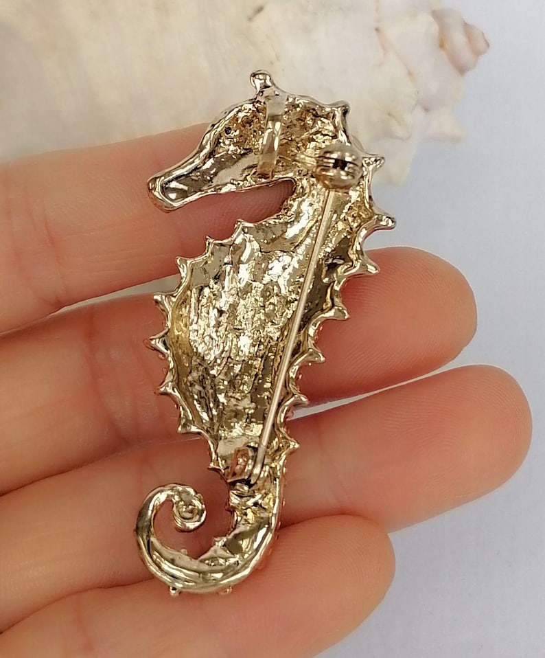 May include: A gold-toned seahorse brooch with a textured surface and a pin closure. The seahorse has a detailed design, including a curled tail and spiky dorsal fin. The brooch is likely made of metal and is a decorative accessory.
