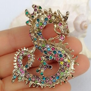 Puede incluir: Broche de drag&oacute;n dorado con brillantes de colores. El drag&oacute;n est&aacute; en una pose estilizada, con su cuerpo formando un bucle. Los brillantes son de varios colores, incluyendo rosa, azul, verde y amarillo. El broche tiene un cierre de alfiler en la parte posterior.