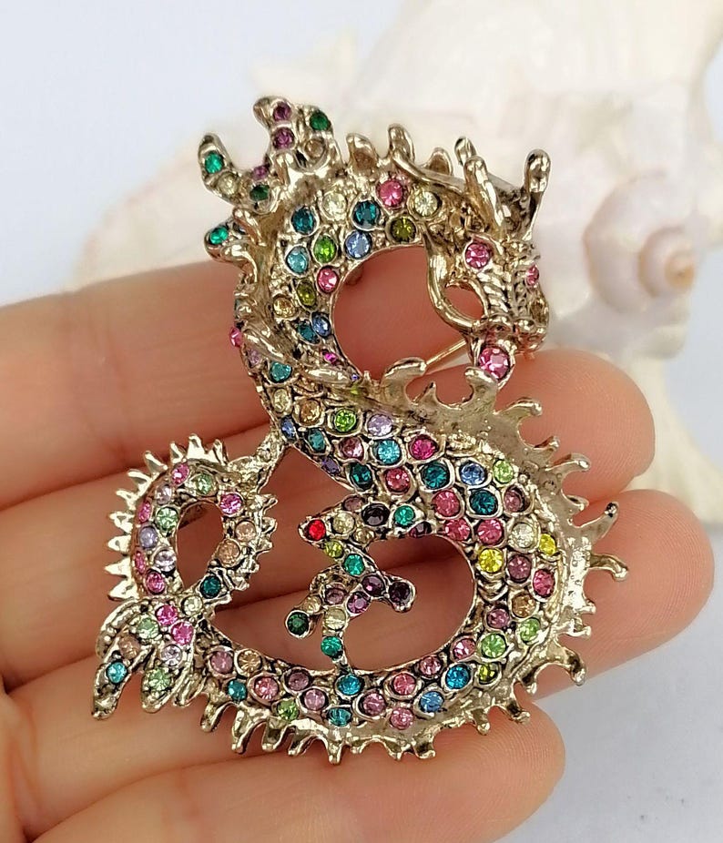 Puede incluir: Broche de drag&oacute;n dorado con brillantes de colores. El drag&oacute;n est&aacute; en una pose estilizada, con su cuerpo formando un bucle. Los brillantes son de varios colores, incluyendo rosa, azul, verde y amarillo. El broche tiene un cierre de alfiler en la parte posterior.