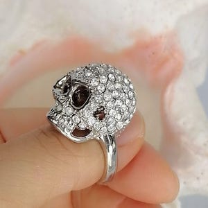 Op de afbeelding: Een zilverkleurige ring met een driedimensionaal schedelontwerp. Het gehele oppervlak van de schedel is ingelegd met talloze kleine, heldere, ronde, gefacetteerde steentjes die het licht reflecteren voor een schitterende uitstraling.