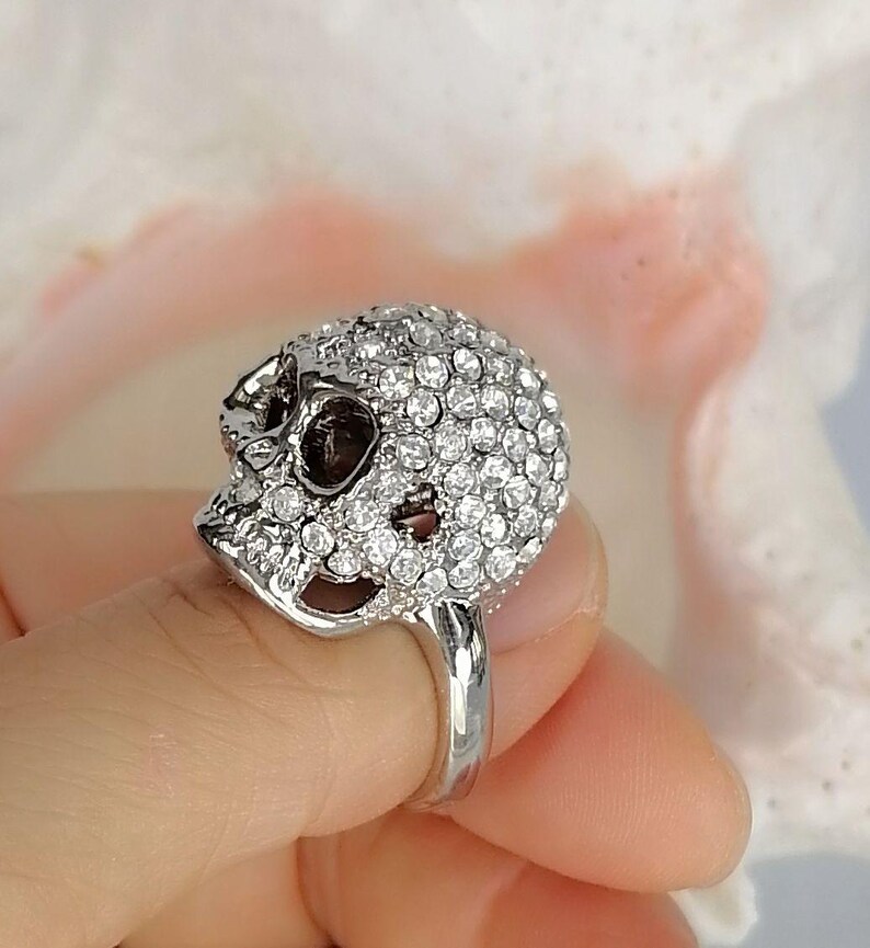 Op de afbeelding: Een zilverkleurige ring met een driedimensionaal schedelontwerp. Het gehele oppervlak van de schedel is ingelegd met talloze kleine, heldere, ronde, gefacetteerde steentjes die het licht reflecteren voor een schitterende uitstraling.