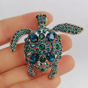 以下が含まれることがあります： 金色のカメのブローチ。ターコイズとグリーンのクリスタルアクセントが施されています。ブローチは、甲羅、頭、ヒレを形成するクリスタルを使用した詳細なデザインが特徴です。クリスタルはさまざまな形とサイズです。