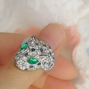 May include: A silver-toned ring shaped like a panther head, featuring two marquise-cut green stones for eyes, numerous small clear white crystals covering the surface, and scattered dark blue accent stones across the metal feline head design.