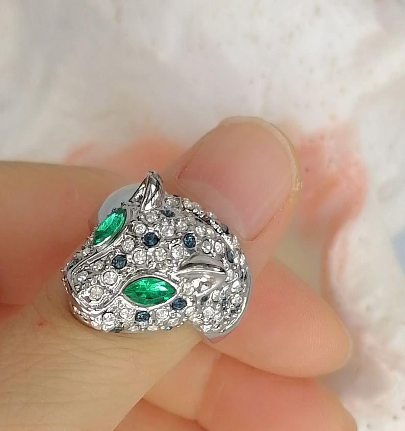 May include: A silver-toned ring shaped like a panther head, featuring two marquise-cut green stones for eyes, numerous small clear white crystals covering the surface, and scattered dark blue accent stones across the metal feline head design.