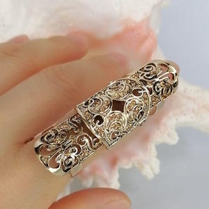 May include: A gold-toned full-finger ring featuring an intricate filigree metalwork design. The surface of the elongated, hinged armour-style ring is embellished with numerous small, sparkling champagne-coloured crystals set within the metal patterns.