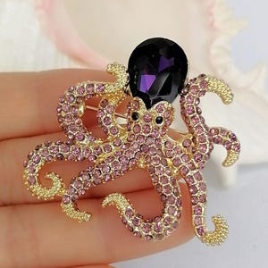 K&ouml;nnte beinhalten: Goldfarbene Oktopus-Brosche mit rosa Strasssteinen und einem gro&szlig;en, dunkelvioletten Edelstein. Die Brosche hat detaillierte Tentakel und schwarze Augen und ist ein funkelndes, auff&auml;lliges Accessoire.