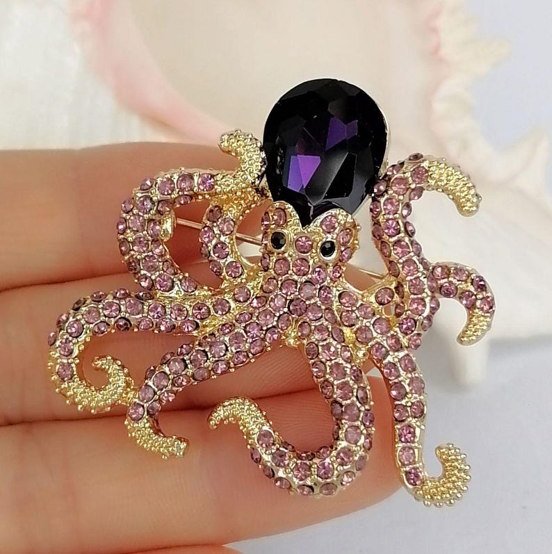 K&ouml;nnte beinhalten: Goldfarbene Oktopus-Brosche mit rosa Strasssteinen und einem gro&szlig;en, dunkelvioletten Edelstein. Die Brosche hat detaillierte Tentakel und schwarze Augen und ist ein funkelndes, auff&auml;lliges Accessoire.