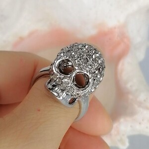 Op de afbeelding: Een zilverkleurige ring met een schedelvormig middendeel. De schedel is bedekt met talloze kleine, heldere, fonkelende steentjes die het gehele oppervlak bedekken, inclusief de oogkassen en de neusholte, wat zorgt voor een textuurrijk uiterlijk.