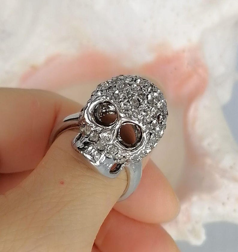 Op de afbeelding: Een zilverkleurige ring met een schedelvormig middendeel. De schedel is bedekt met talloze kleine, heldere, fonkelende steentjes die het gehele oppervlak bedekken, inclusief de oogkassen en de neusholte, wat zorgt voor een textuurrijk uiterlijk.