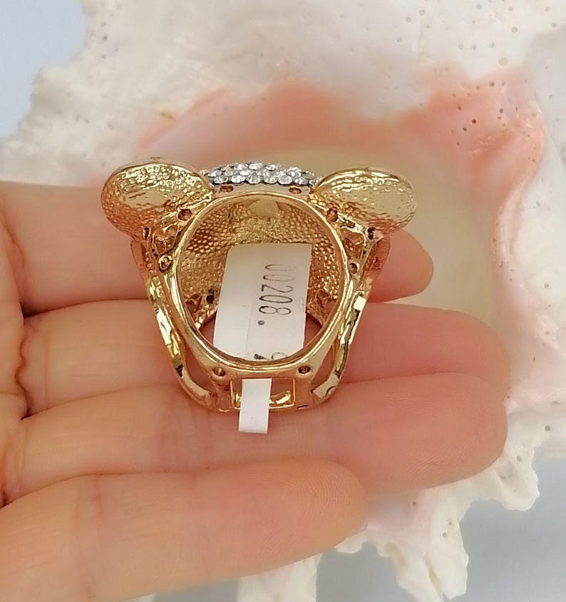 May include: A gold-toned ring featuring a bear-shaped head adorned with clear crystals. The metal surface has a textured finish, and a white rectangular tag with the printed text 00208 is attached to the inner band of the ring.
