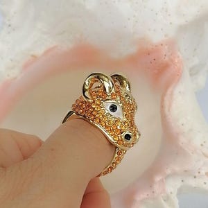May include: A gold-toned ring shaped like a ram's head, featuring a surface covered in small, sparkling orange crystals. The ram's eyes are accented with white enamel and black centres, while the horns and snout are polished gold-tone metal.