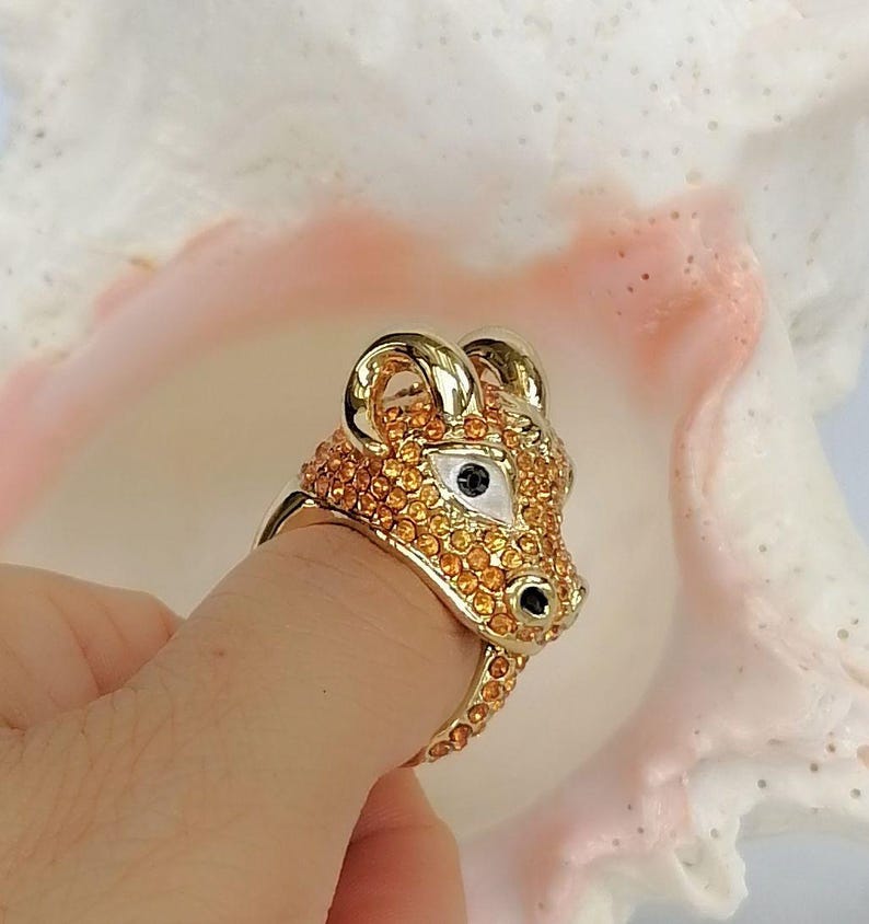 May include: A gold-toned ring shaped like a ram's head, featuring a surface covered in small, sparkling orange crystals. The ram's eyes are accented with white enamel and black centres, while the horns and snout are polished gold-tone metal.