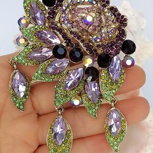 Puede incluir: Un broche con forma de flor que presenta un dise&ntilde;o de rosa central incrustado con cristales morados e iridiscentes. La pieza incluye detalles en forma de hoja pavimentados con peque&ntilde;os cristales verdes, piedras lavanda de corte marquesa y tres colgantes.