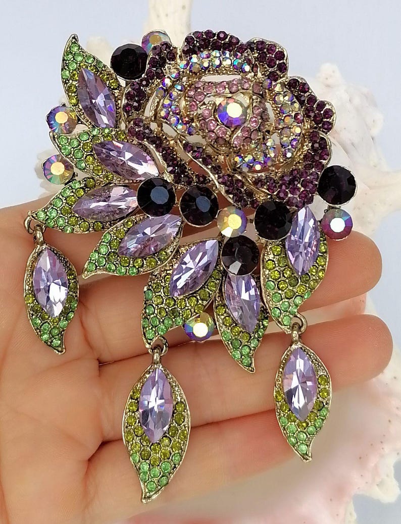 Puede incluir: Un broche con forma de flor que presenta un dise&ntilde;o de rosa central incrustado con cristales morados e iridiscentes. La pieza incluye detalles en forma de hoja pavimentados con peque&ntilde;os cristales verdes, piedras lavanda de corte marquesa y tres colgantes.