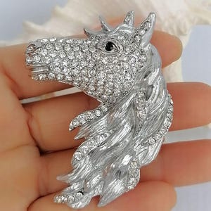 K&ouml;nnte beinhalten: Silberfarbene Pferdekopfbrosche, besetzt mit klaren Kristallen. Das Auge des Pferdes ist ein kleiner schwarzer Stein. M&auml;hne und Kopf sind mit strukturiertem Metall detailliert. Ein dekoratives Accessoire.