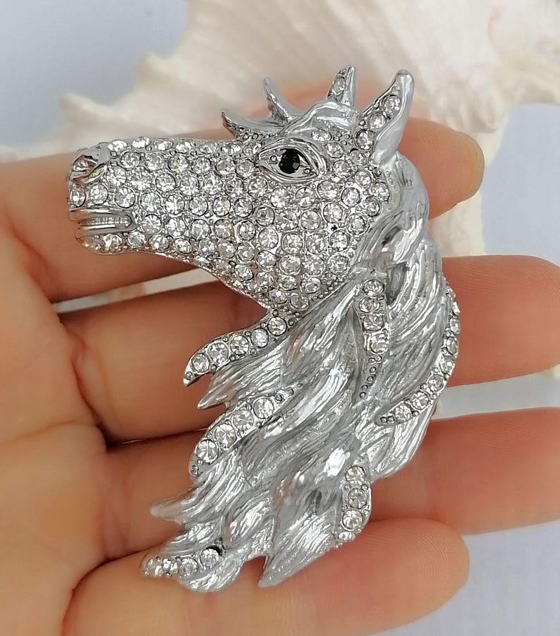 K&ouml;nnte beinhalten: Silberfarbene Pferdekopfbrosche, besetzt mit klaren Kristallen. Das Auge des Pferdes ist ein kleiner schwarzer Stein. M&auml;hne und Kopf sind mit strukturiertem Metall detailliert. Ein dekoratives Accessoire.