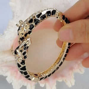 May include: A hinged bangle bracelet featuring a gold-tone metal finish with black enamel giraffe-print spots. The clasp area is designed as two giraffe heads facing each other, accented with small clear crystals along the top edge of the band.