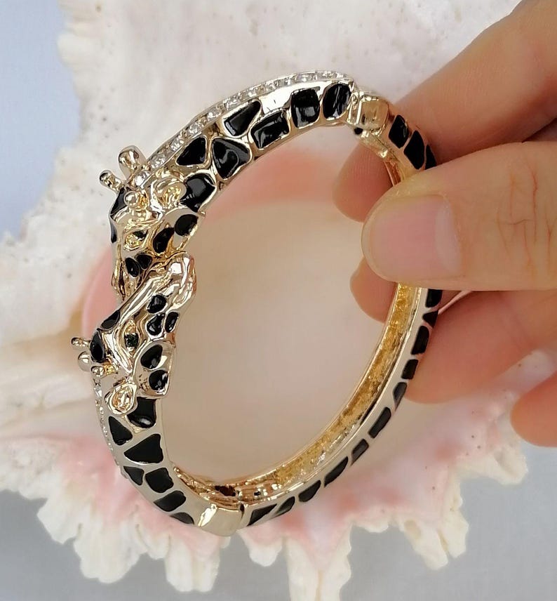 May include: A hinged bangle bracelet featuring a gold-tone metal finish with black enamel giraffe-print spots. The clasp area is designed as two giraffe heads facing each other, accented with small clear crystals along the top edge of the band.