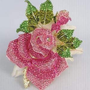 Puede incluir: Broche de rosa enjoyada con cristales rosas y verdes. El broche presenta una rosa grande y abierta con p&eacute;talos detallados y capullos m&aacute;s peque&ntilde;os, rodeada de hojas verdes y verde amarillento. El tallo es dorado.