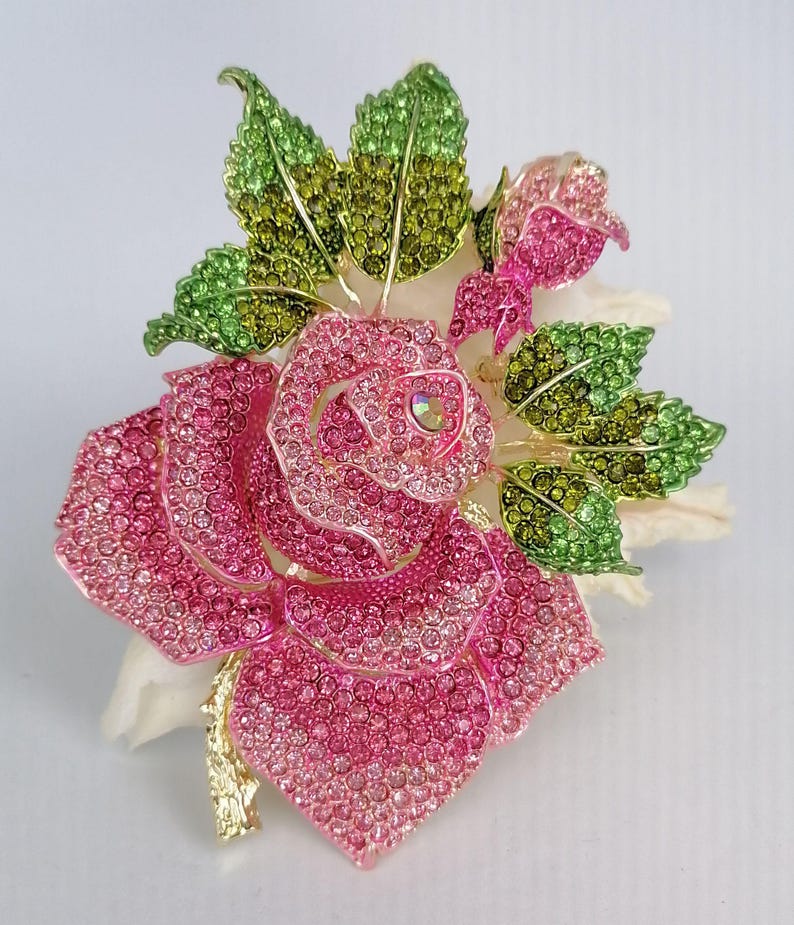 Puede incluir: Broche de rosa enjoyada con cristales rosas y verdes. El broche presenta una rosa grande y abierta con p&eacute;talos detallados y capullos m&aacute;s peque&ntilde;os, rodeada de hojas verdes y verde amarillento. El tallo es dorado.