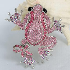 K&ouml;nnte beinhalten: Ein Frosch-Broschen-Anstecker mit silberfarbenem Metallrahmen, besetzt mit rosa Strasssteinen. Der K&ouml;rper des Frosches ist mit dunkleren rosa Steinen umrandet, die Augen sind schwarz. Ein verspieltes Accessoire.