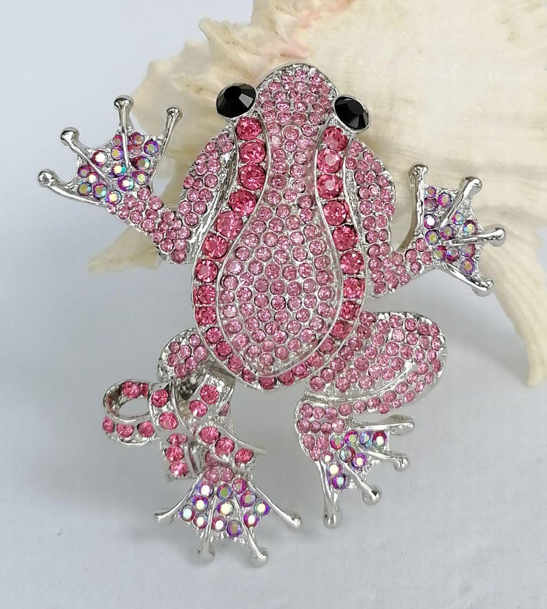 K&ouml;nnte beinhalten: Ein Frosch-Broschen-Anstecker mit silberfarbenem Metallrahmen, besetzt mit rosa Strasssteinen. Der K&ouml;rper des Frosches ist mit dunkleren rosa Steinen umrandet, die Augen sind schwarz. Ein verspieltes Accessoire.