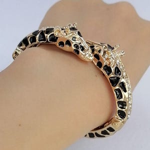 May include: A gold-tone hinged bangle bracelet featuring two giraffe heads meeting at the center. The bracelet is decorated with black enamel spots and clear crystals along the sides, mimicking the pattern of a giraffe's coat in a metallic finish.
