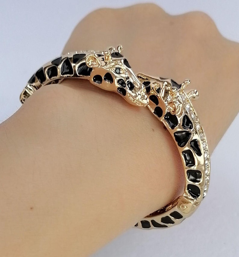 May include: A gold-tone hinged bangle bracelet featuring two giraffe heads meeting at the center. The bracelet is decorated with black enamel spots and clear crystals along the sides, mimicking the pattern of a giraffe's coat in a metallic finish.