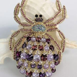 May include: A gold-coloured spider brooch, embellished with purple and pink rhinestones. The brooch has a detailed design, with a central cluster of purple gems and a body adorned with various shades of purple and pink crystals. The spider's legs are embellished with pink rhinestones.