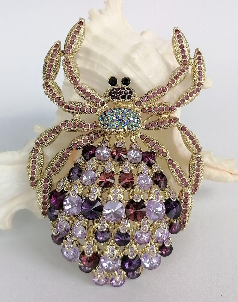 May include: A gold-coloured spider brooch, embellished with purple and pink rhinestones. The brooch has a detailed design, with a central cluster of purple gems and a body adorned with various shades of purple and pink crystals. The spider's legs are embellished with pink rhinestones.