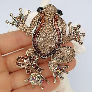 K&ouml;nnte beinhalten: Ein mit Juwelen besetzter Frosch-Broche mit einem goldfarbenen Metallsockel. Der Frosch ist mit kleinen, funkelnden Kristallen in Braun-, Bernstein- und schillernden Farbt&ouml;nen bedeckt. Die Augen sind schwarz. Das Schmuckst&uuml;ck ist ein dekoratives Accessoire.