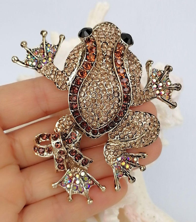 K&ouml;nnte beinhalten: Ein mit Juwelen besetzter Frosch-Broche mit einem goldfarbenen Metallsockel. Der Frosch ist mit kleinen, funkelnden Kristallen in Braun-, Bernstein- und schillernden Farbt&ouml;nen bedeckt. Die Augen sind schwarz. Das Schmuckst&uuml;ck ist ein dekoratives Accessoire.
