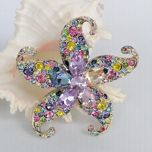 May include: A flower-shaped brooch with a gold-tone metal base, encrusted with a variety of colorful rhinestones. The brooch features a central cluster of large, teardrop-shaped stones in shades of purple and peach, surrounded by smaller, round rhinestones in various colors.
