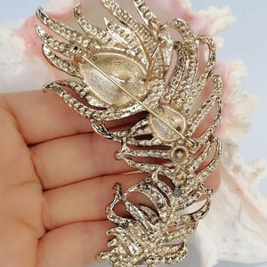 May include: A gold-toned metal brooch shaped like a large, detailed feather. The back of the brooch features a horizontal pin mechanism with a safety clasp. The textured metal surface shows intricate vein patterns throughout the entire feather design.