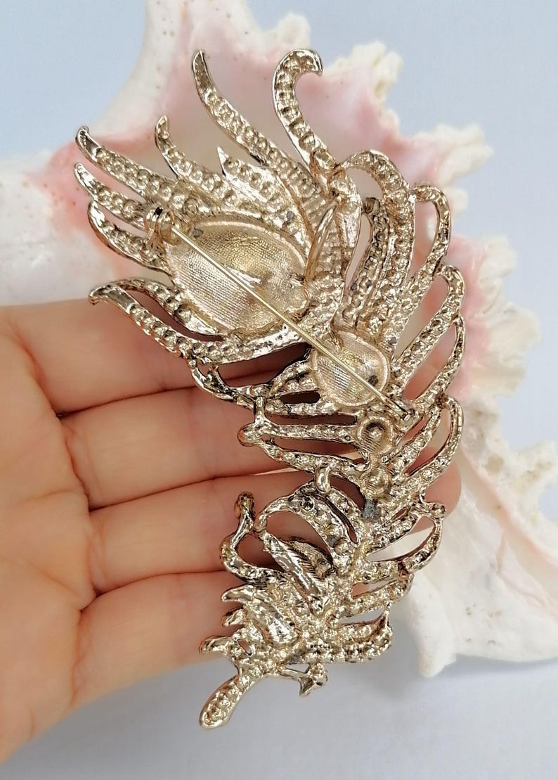 May include: A gold-toned metal brooch shaped like a large, detailed feather. The back of the brooch features a horizontal pin mechanism with a safety clasp. The textured metal surface shows intricate vein patterns throughout the entire feather design.