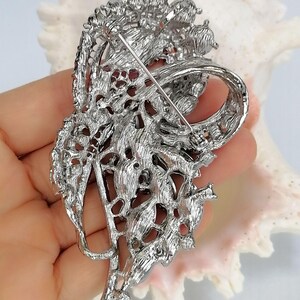 May include: A silver-toned metal brooch featuring an intricate, openwork floral design with a textured, filigree-like finish. The back of the piece displays a long, thin horizontal pin mechanism with a secure clasp for attachment to garments.
