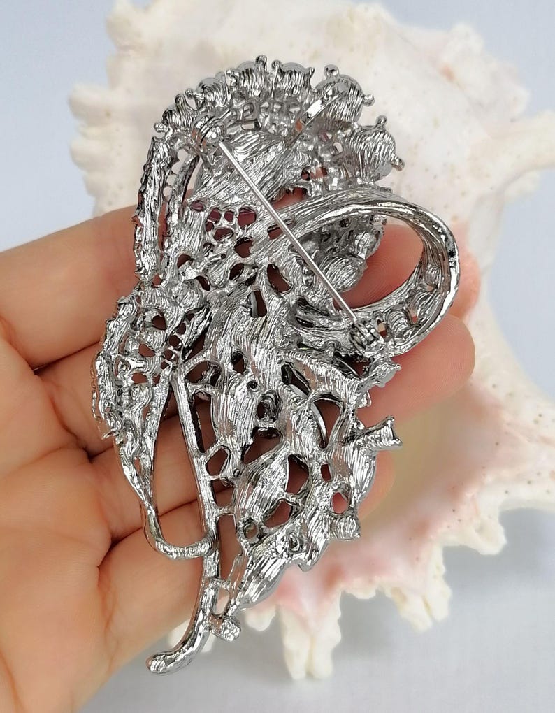 May include: A silver-toned metal brooch featuring an intricate, openwork floral design with a textured, filigree-like finish. The back of the piece displays a long, thin horizontal pin mechanism with a secure clasp for attachment to garments.
